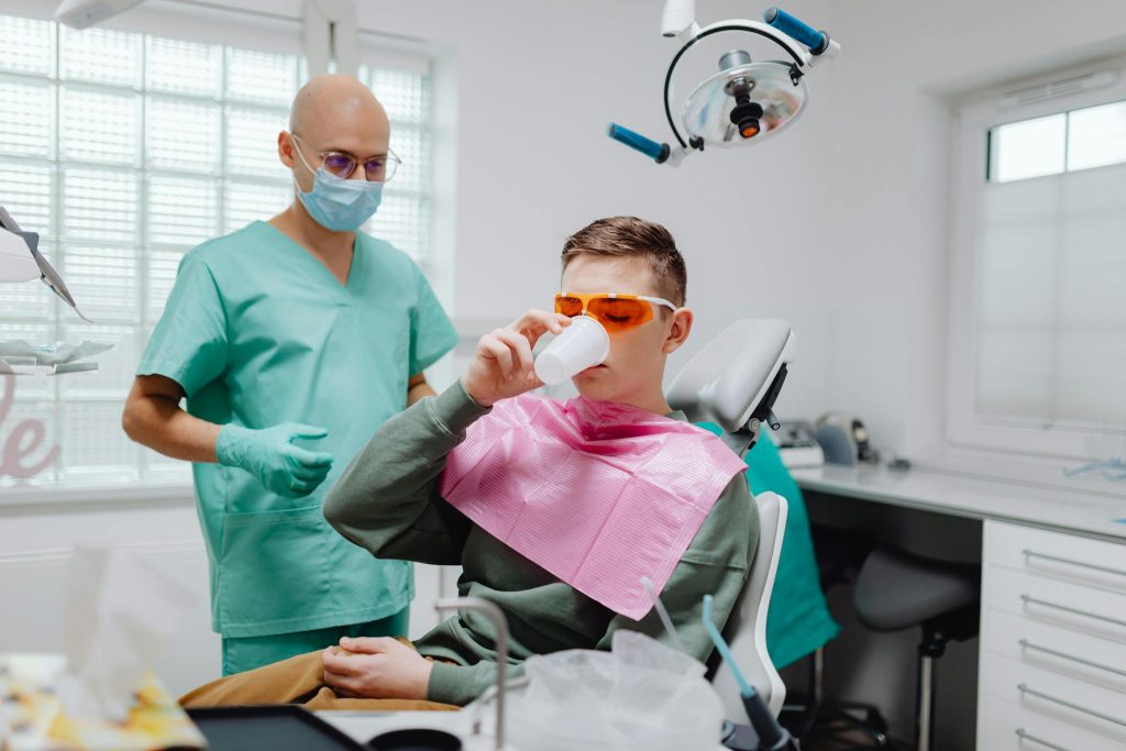 A teenager sits in a dental chair drinking from a cup, attended by a dental professional.